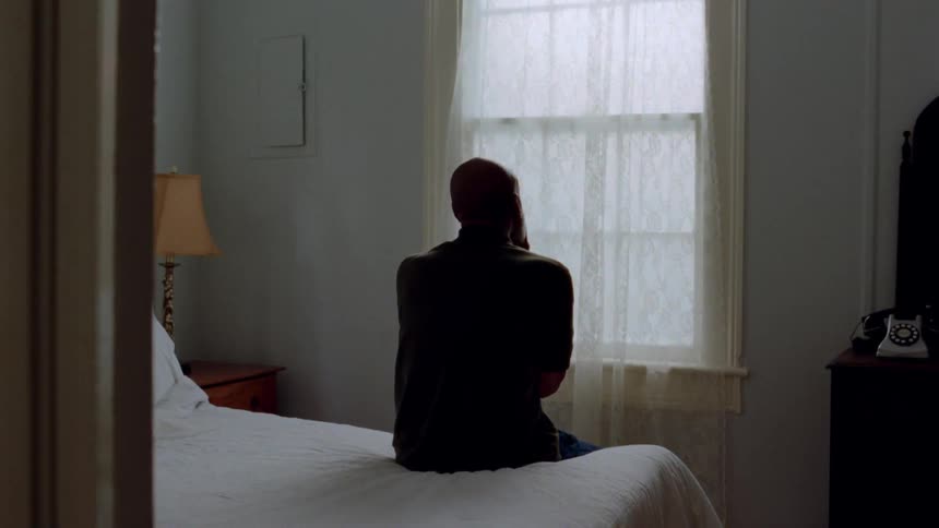 Man sits at the edge of a bed rubbing his face with his hands