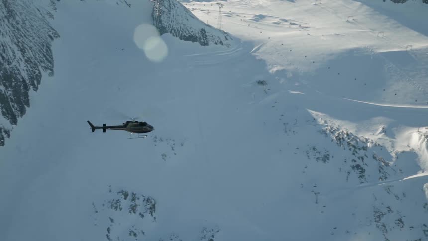 A helicopter flies above a snow covered mountain range during the day