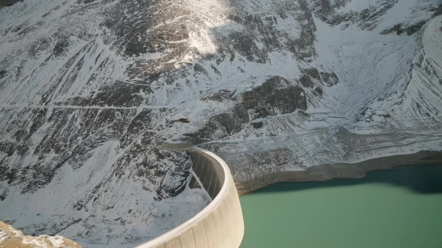 A helicopter flies over a dam near snowy mountains
