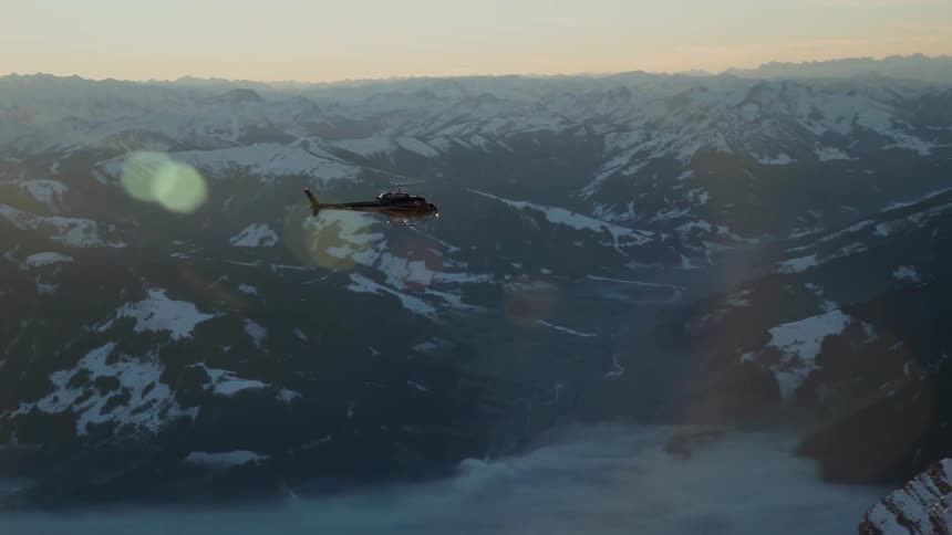 A helicopter flies over snowy mountains at sunset