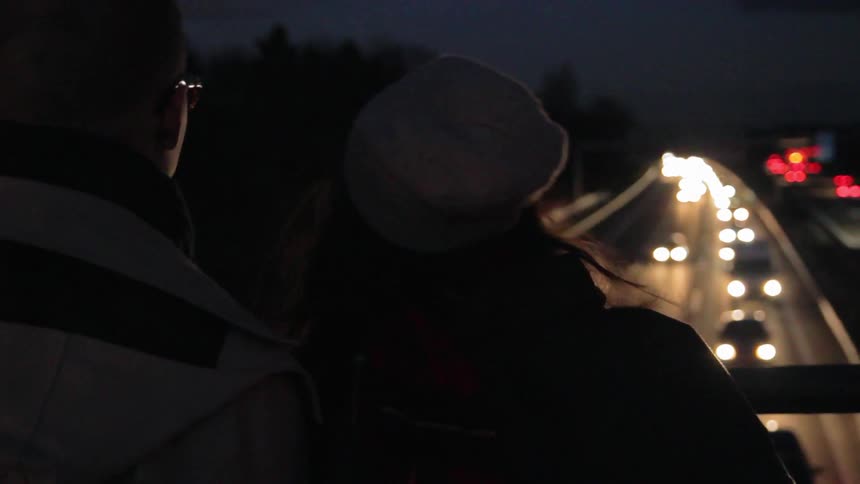 A couple holds hands while standing on an overpass above the highway