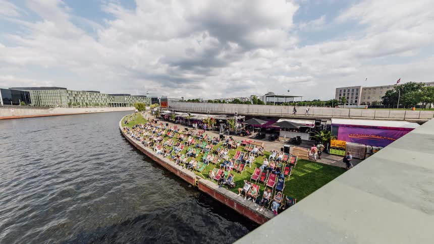 Crowd of people sit on lawn chairs next to a river of water as boats pass by