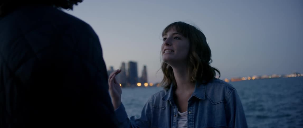 Man and woman talk outside by a river with a city across the river