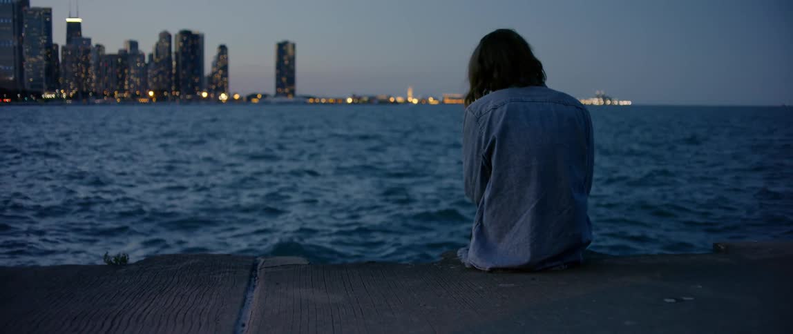 A person sits on the edge of a pier looking across at a city skyline
