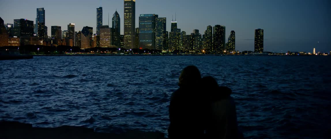 Man and woman cuddle while sitting next to city river at dusk