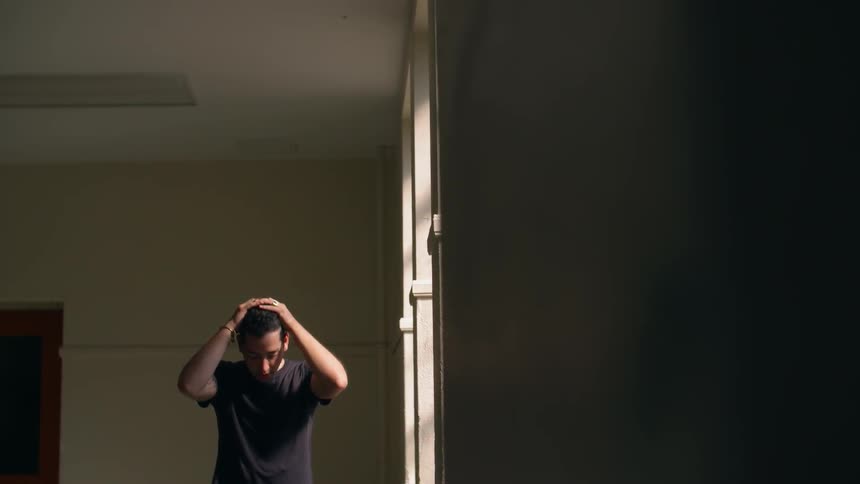 Man standing in dark room by the window rubbing hands through his hair