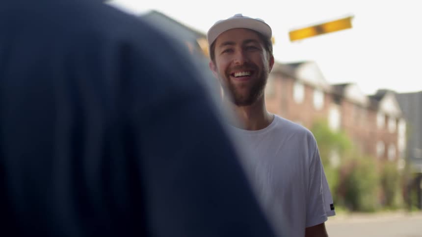 Man smiles and talks to another person outside on the street next to an apartment building