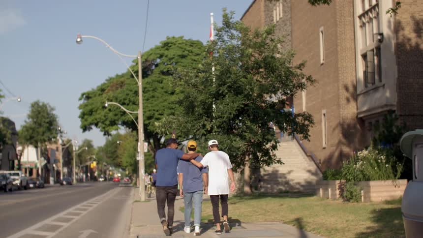 A group of friends are walking with their arms on each others shoulders down the sidewalk