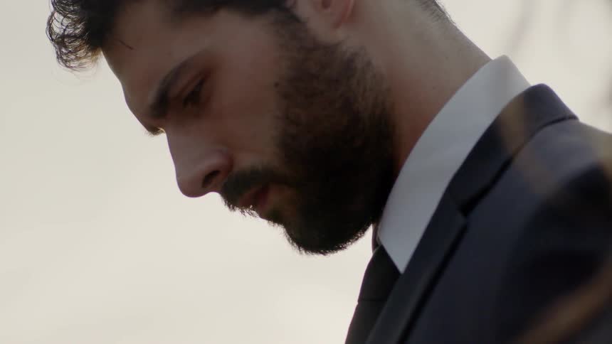 Bearded man in suit looks down with solemn expression