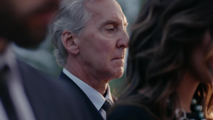 An older man is standing behind some other people looking sad