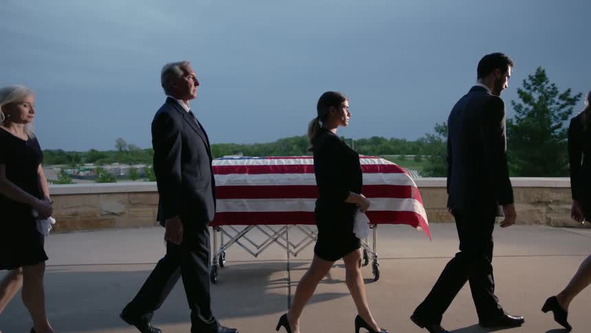 Grieving people walk past a flag draped casket