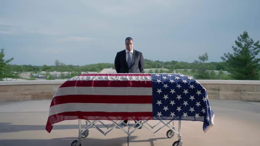 A man is standing in front of a american flag covered coffin