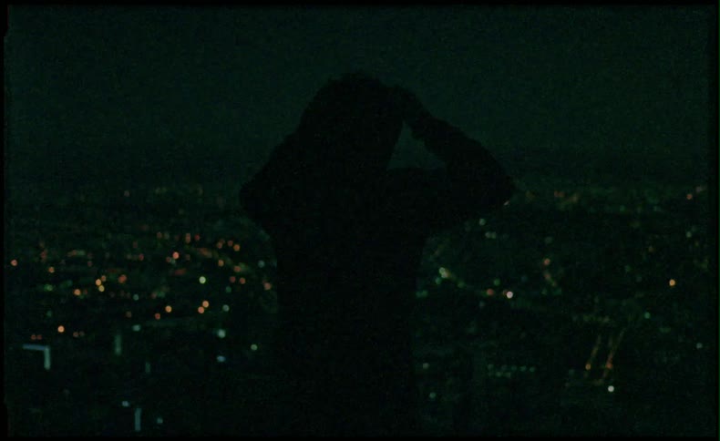 A man stands atop a cliff overlooking the city at night