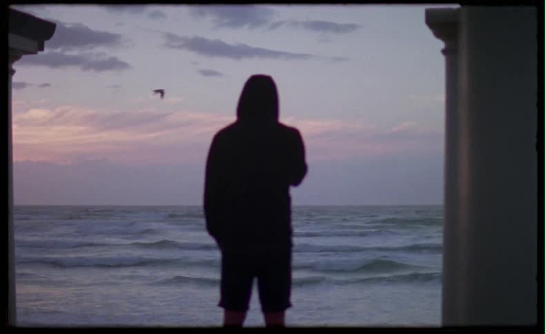 Person in hoodie smoking while looking at beach on sunset