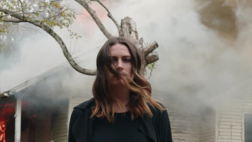 A woman stands in front of a smoking and burning house