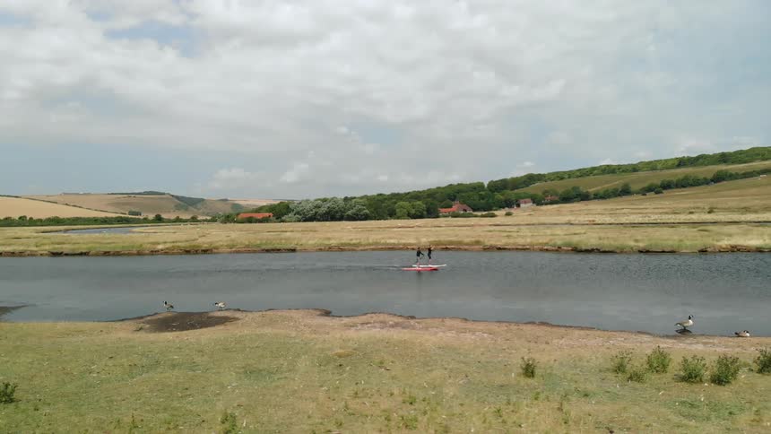 People paddle a raft along a river as sheep graze on nearby grass