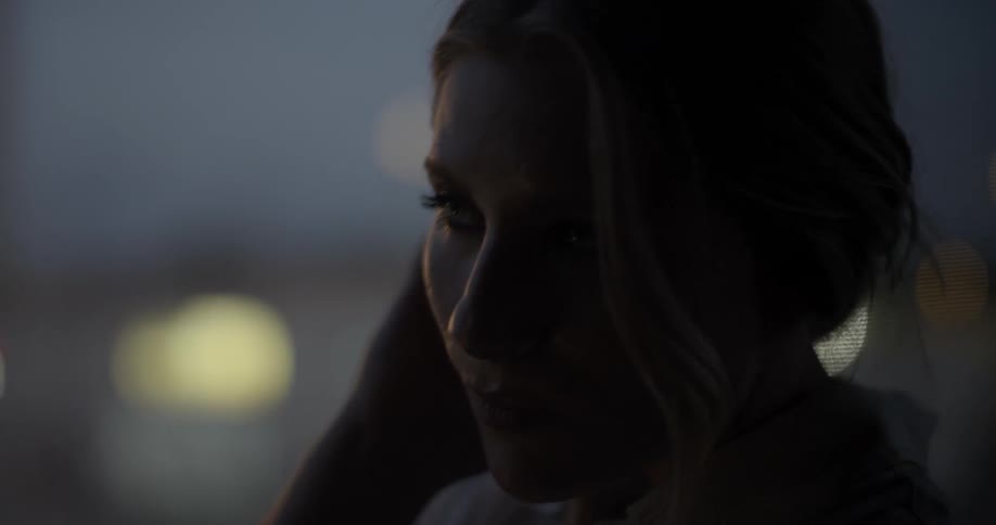 A woman brushes her hair back and hold her arm tight as she stands near a window at dusk