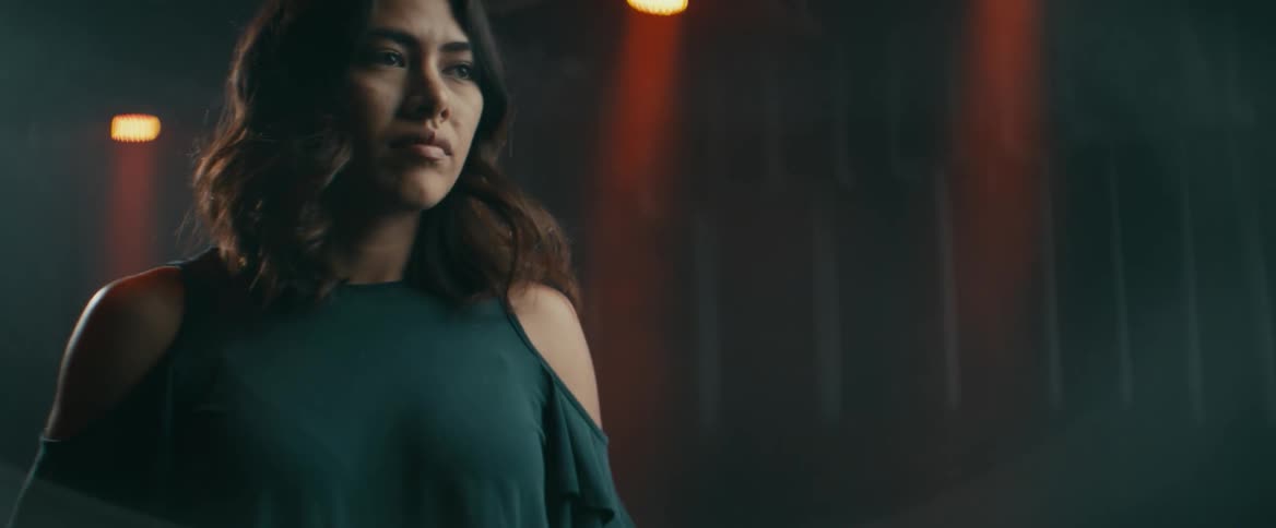 A woman walking in a dimly lit studio stops to take a deep breath and compose herself