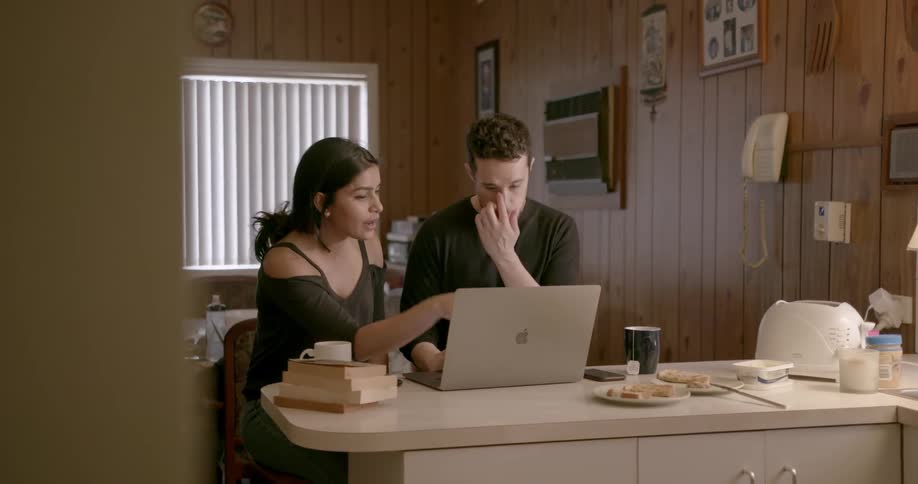 A man and woman are discussing something on a laptop as they sit at their kitchen counter