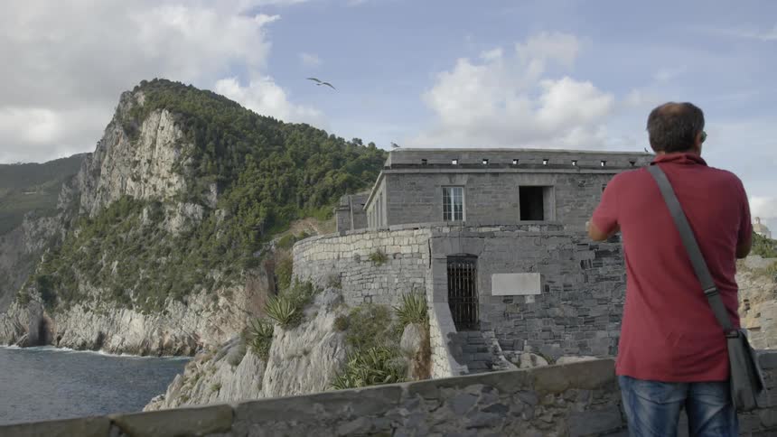 Man taking pictures of an ancient structure that is seaside on a cloudy day