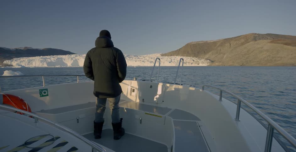 A man is standing on the prow of a boat as it approaches a glacier