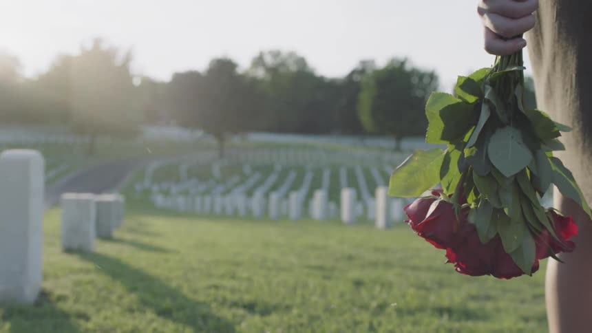 A woman in a dress is walking in a graveyard with some flowers