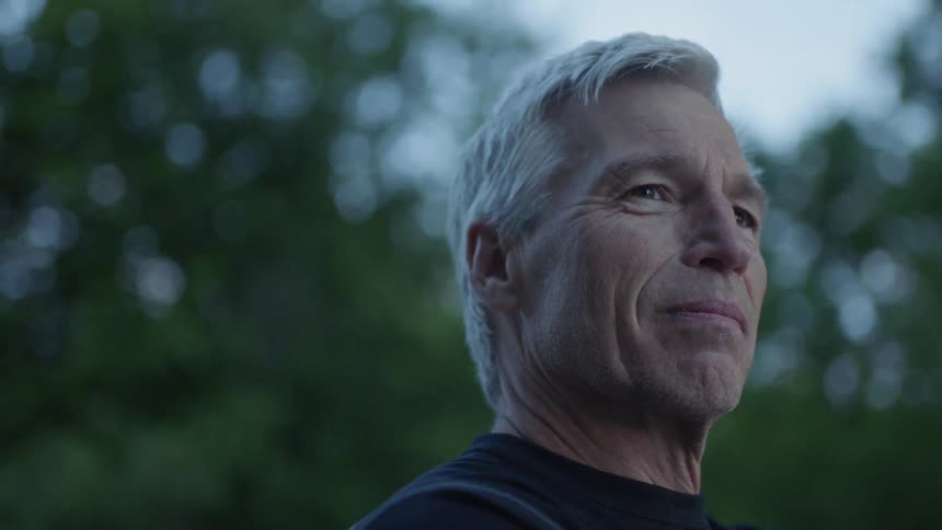 An elderly man smiles while standing near trees