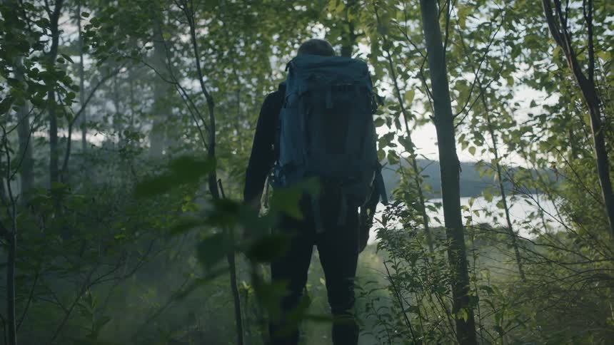 Man wearing backpack is walking through trees towards lake as sun sets
