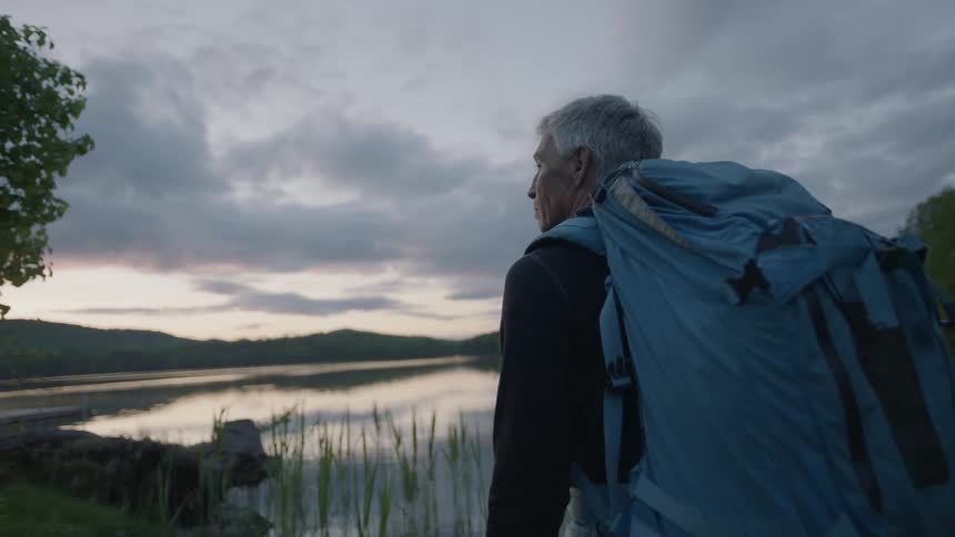 A man with grey hair and a backpack is standing in front of a lake