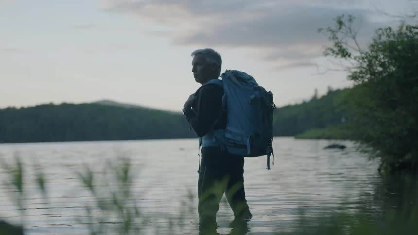 A man carrying a backpack stands knee deep in a lake
