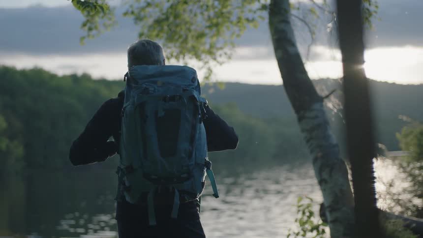 Man with backpack stands looking at a lake at sunset