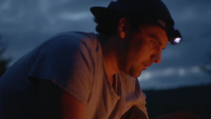 A man stands over a campfire with a headlamp on his head