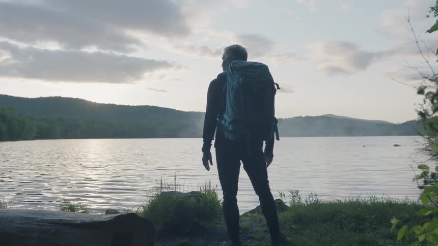 A man with a backpack is standing next to a log and a river