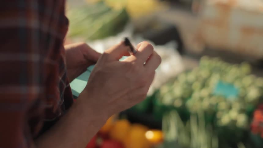 A person writes something on a blue note pad while standing next to a vegetable stand