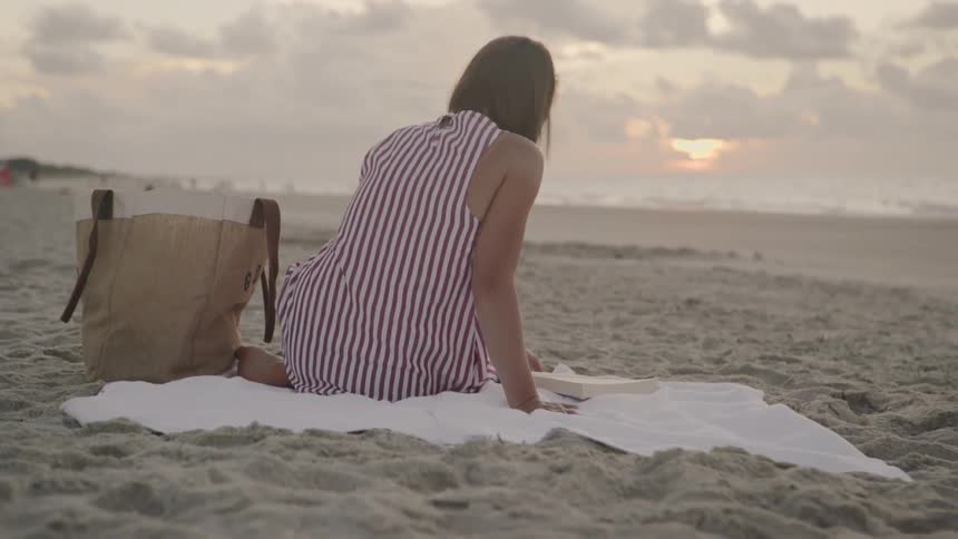 Woman sitting on towel on beach is flipping through book at sunset