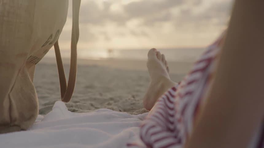 A person lays on the sand watching two others near the water at sunset