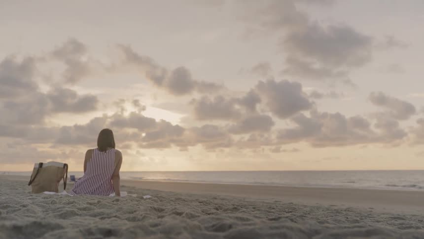 A woman sits on a towel looking out at the ocean at sunrise