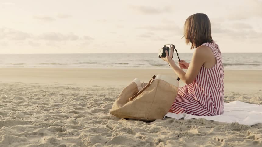 Woman sits on a blanket at the beach and retrieves her camera from a bag