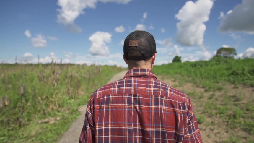 Man in a flannel shirt walking through a field on a partly cloudy day