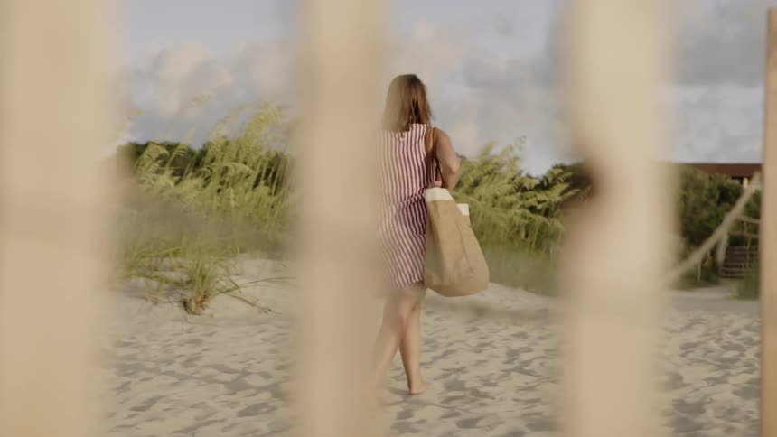 A woman walks on the beach carrying a bag and blanket