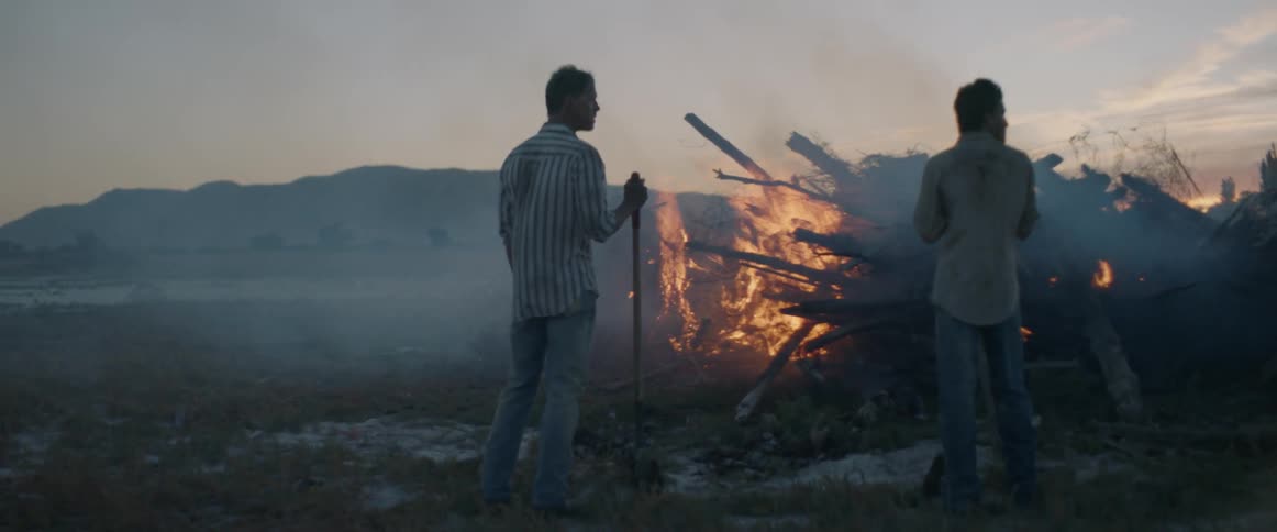 Two men with shovels stand near a fire in a field