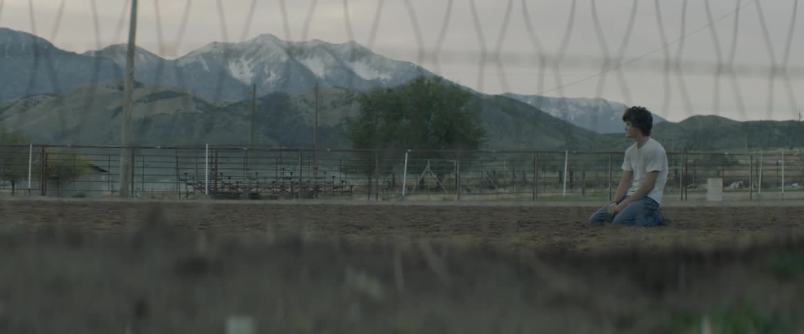 Young man is sitting on knees on dirt inside rodeo ring at dusk