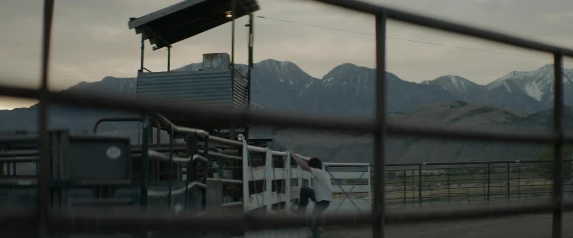 A man jumps up on the fence of the rodeo ring and sits