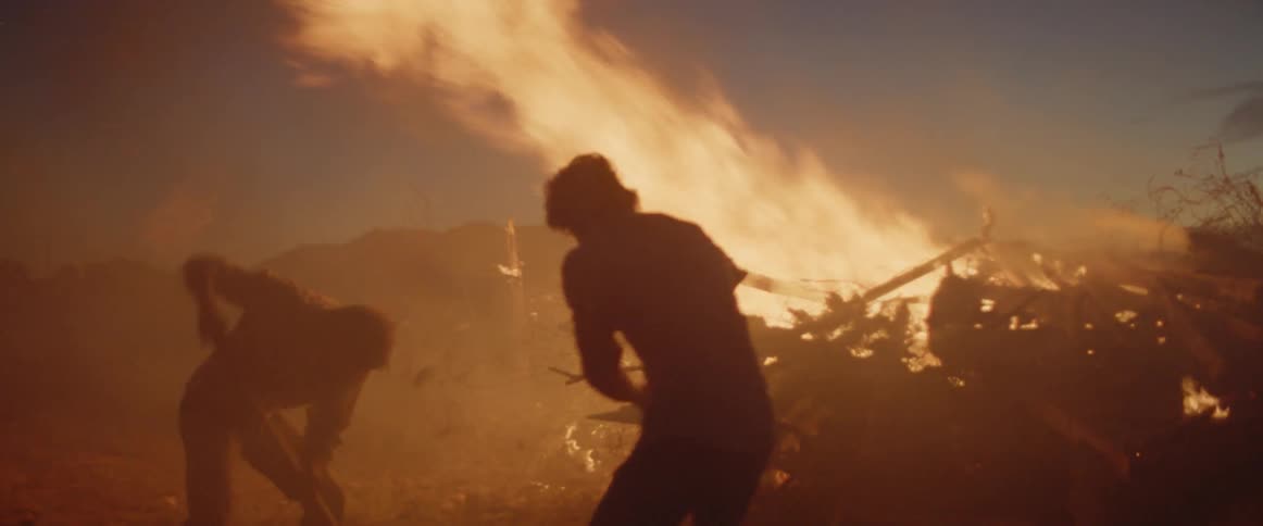 Two men using shovels to fling dirt onto a large bonfire at night