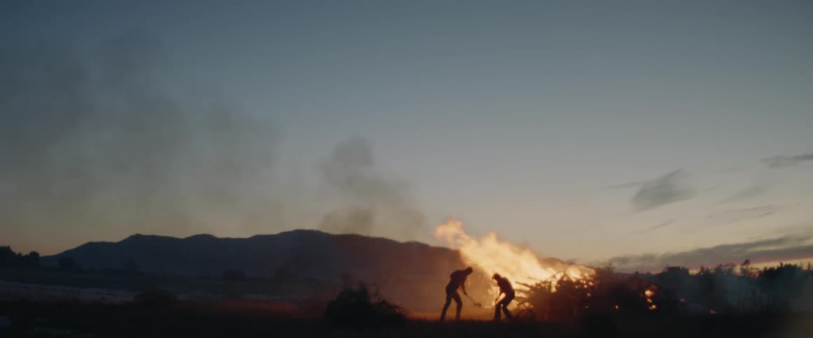 Two people in the distance dig dirt on a large bonfire
