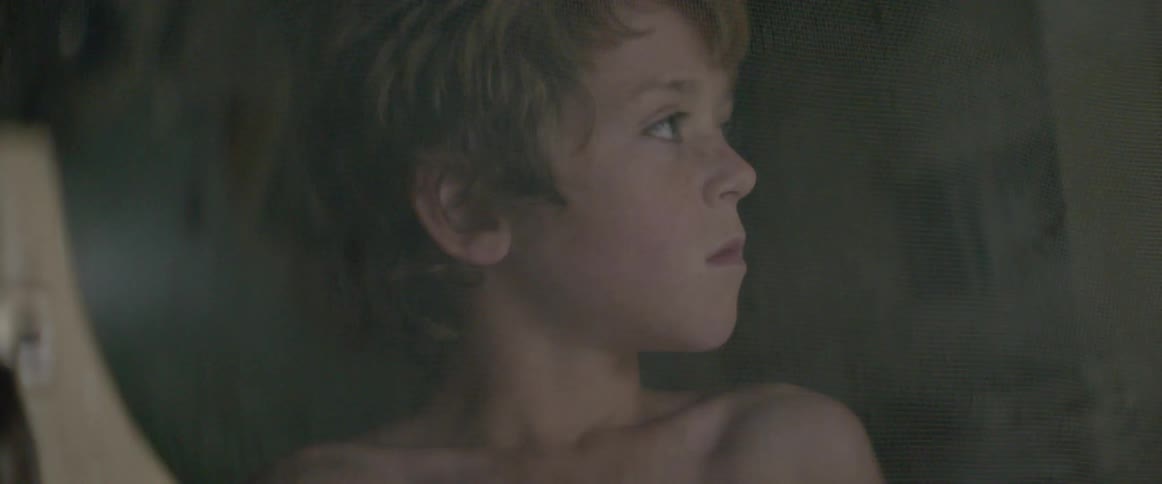 A kid without a shirt is peaking through a screen door