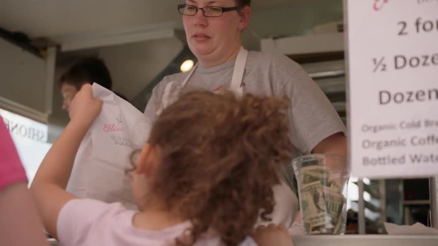 A woman is putting pastries into a white paper sack and handing them to a small child