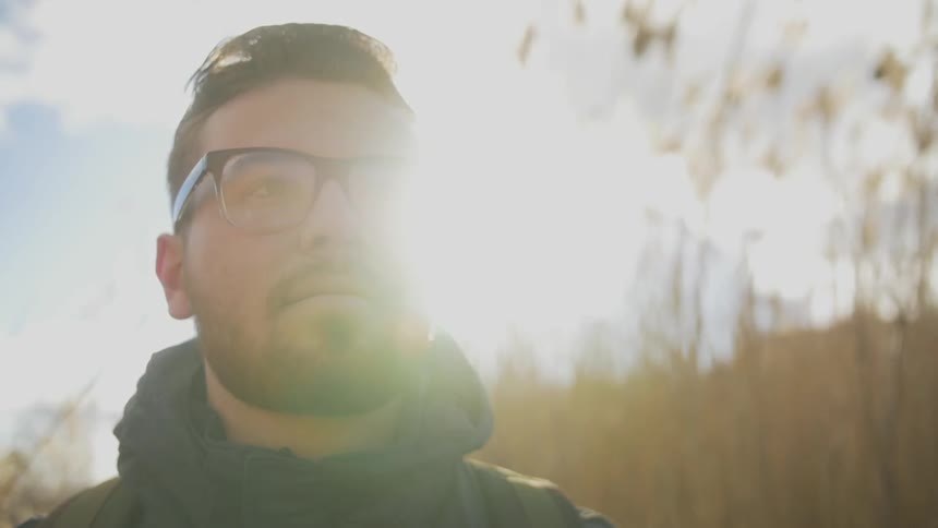 A young, bearded, brunette man wearing glasses standing outdoors looking up at something as the sun glares behind him