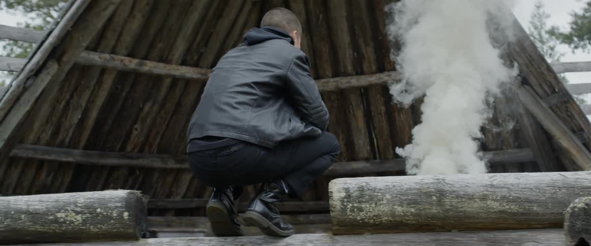 A man crouches down by smoke rising in the hut