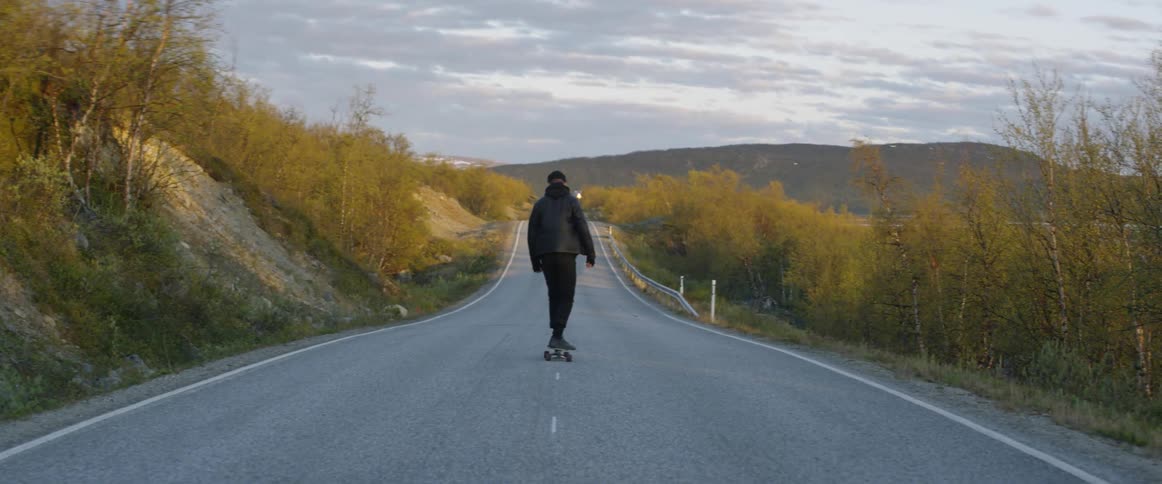 A man rides a skateboard down the center of a rural road